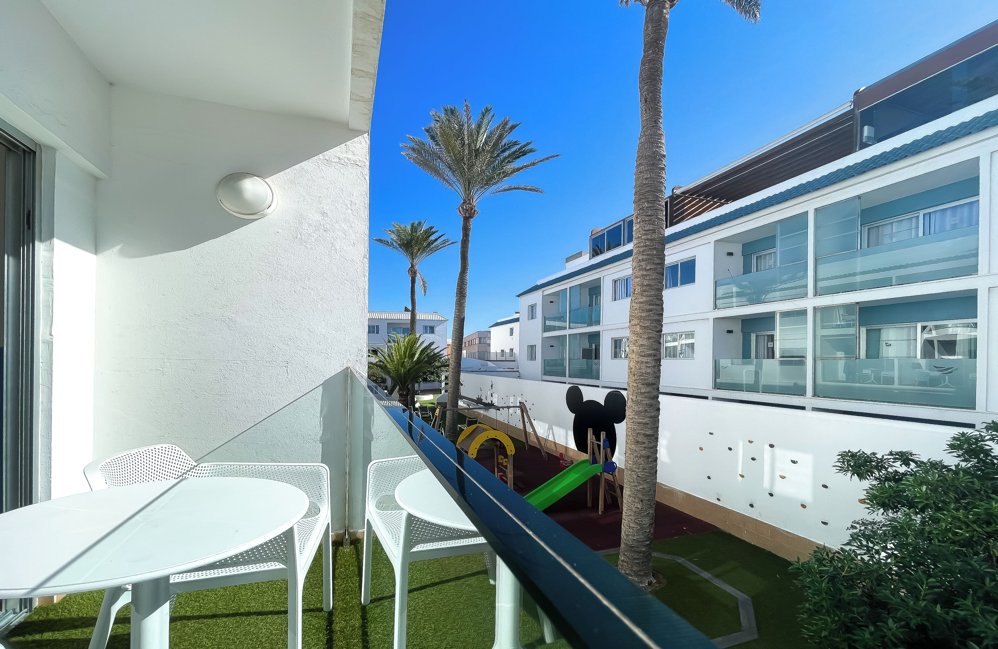 A balcony with a white table and chairs, a children's play area with a slide, and a view of a modern building with large windows.