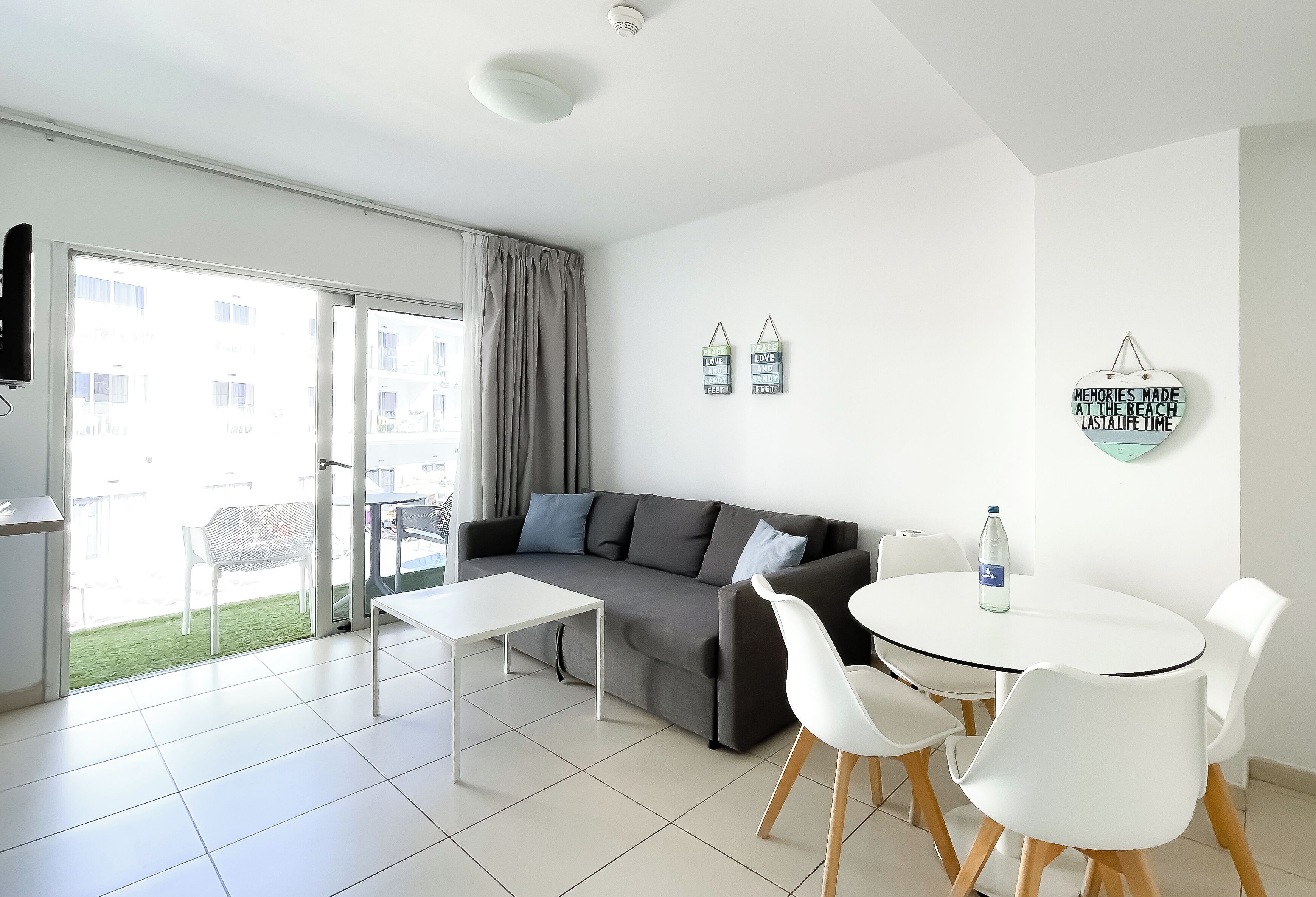 Een moderne woonkamer met een grijze bank, witte stoelen, een kleine tafel en een salontafel met een glazen blad. Er hangen twee decoraties aan de muur en een raam met gordijnen.