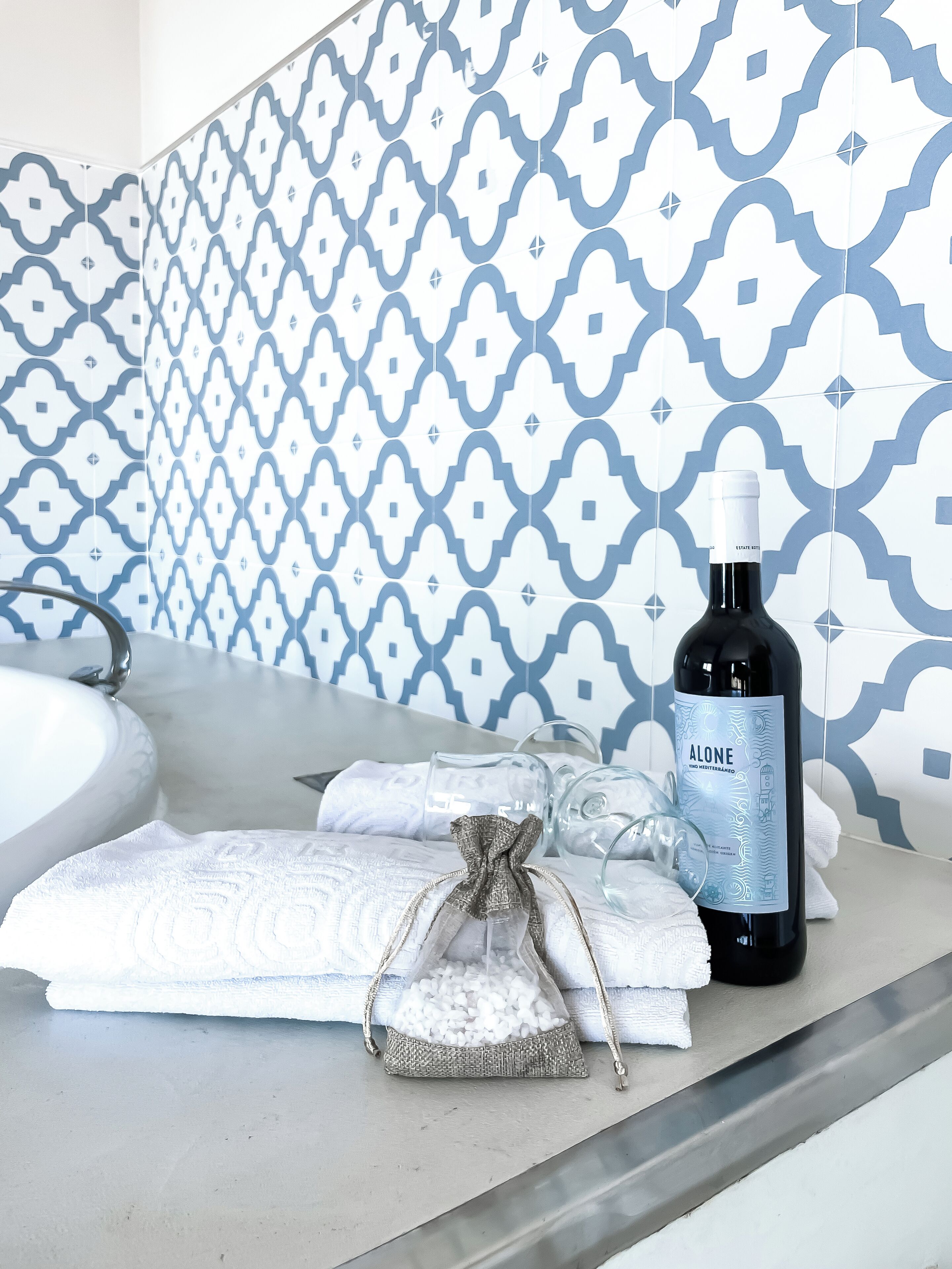 A bathroom with a patterned tile backsplash, a sink, a bottle of wine, towels, and a glass pitcher.