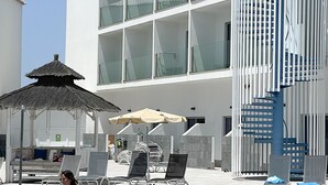 A swimming pool area with lounge chairs, a thatched umbrella, and a modern building with large windows. - Corralejo Surfing Colors Hotel&Apartments (La Oliva)