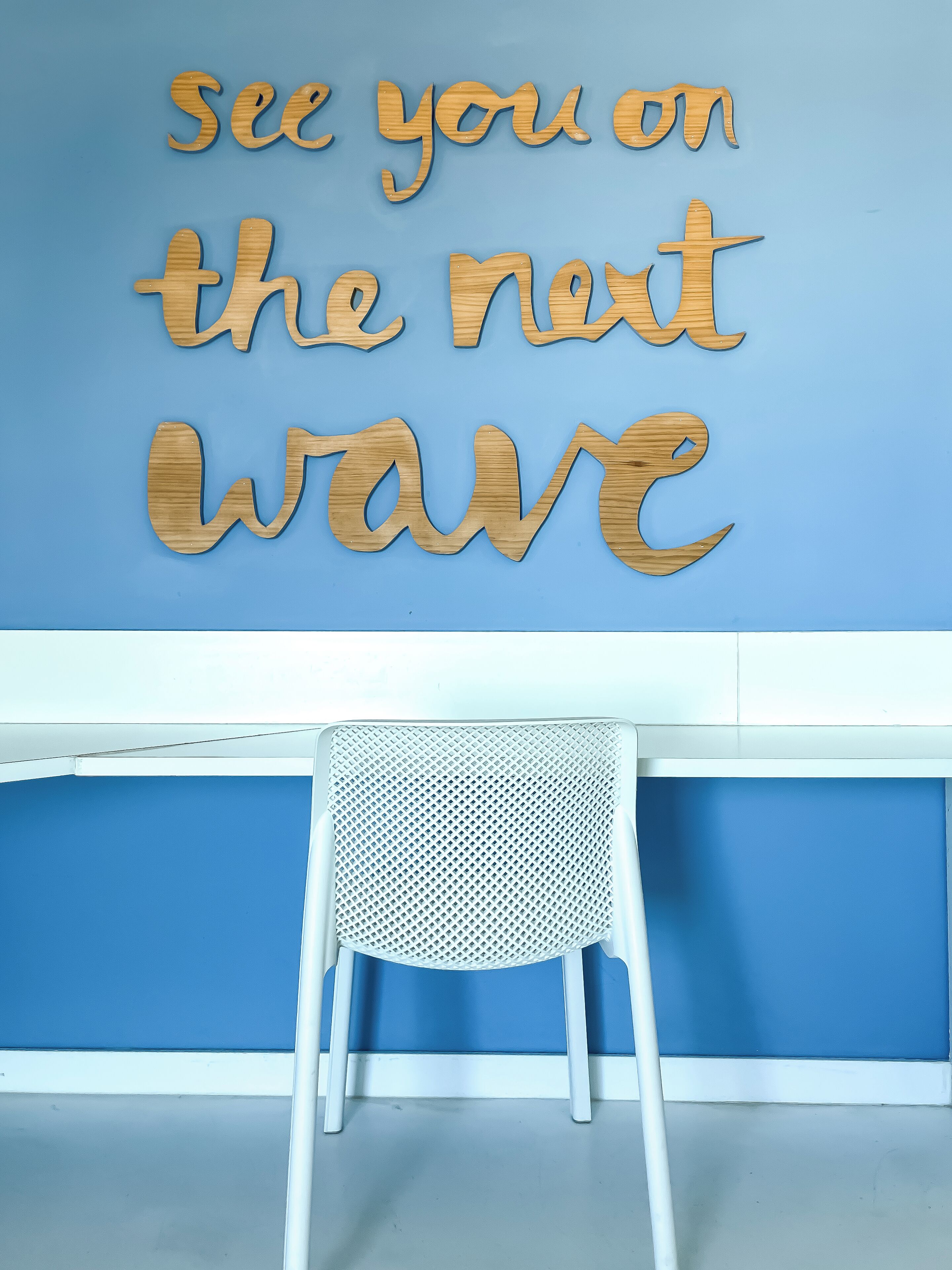 A wall with wooden letters spelling "see you on the next wave" and a white chair in front.
