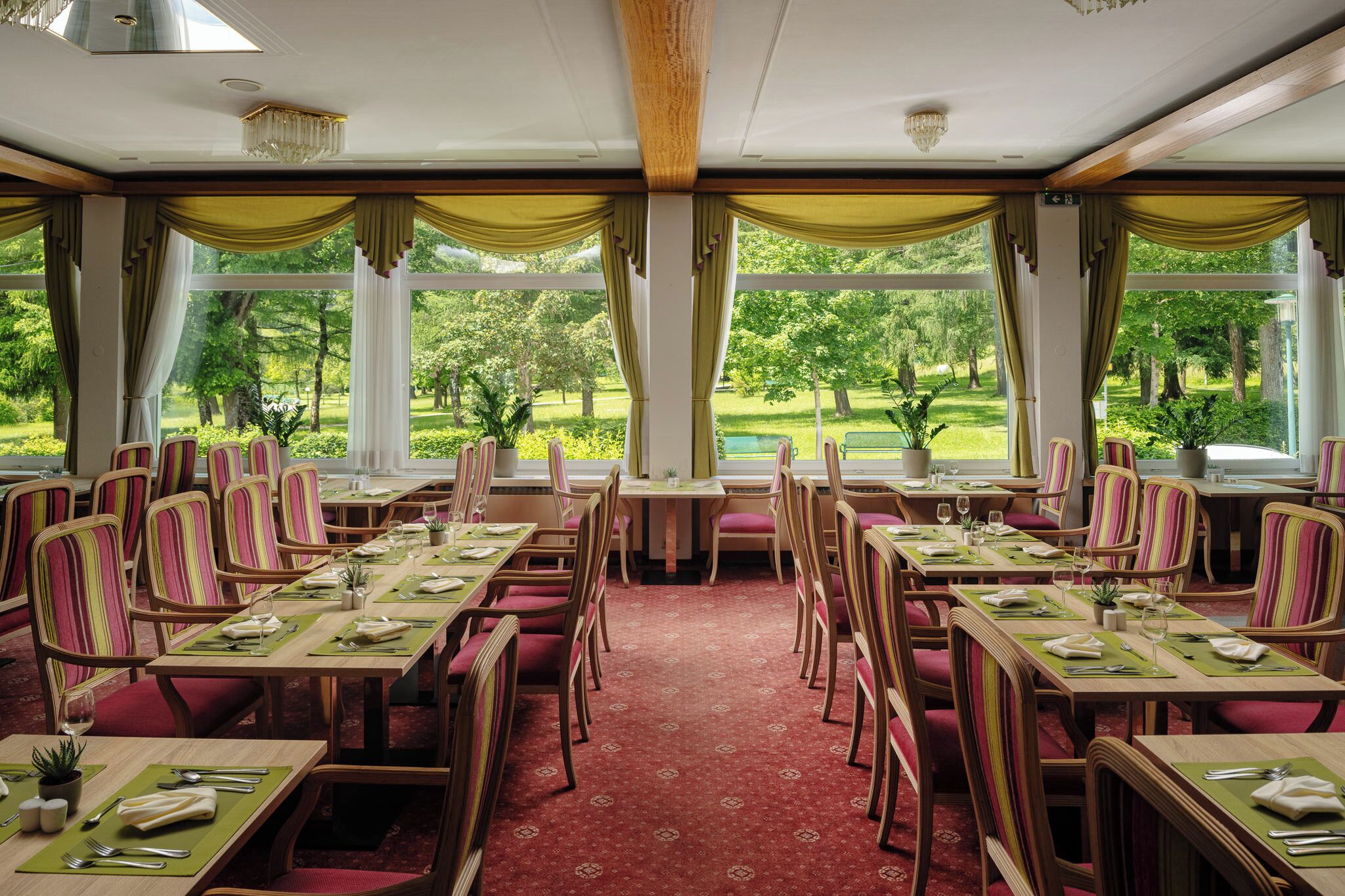 A dining area with tables set for four, chairs with striped upholstery, and a view of a lush garden through large windows.
