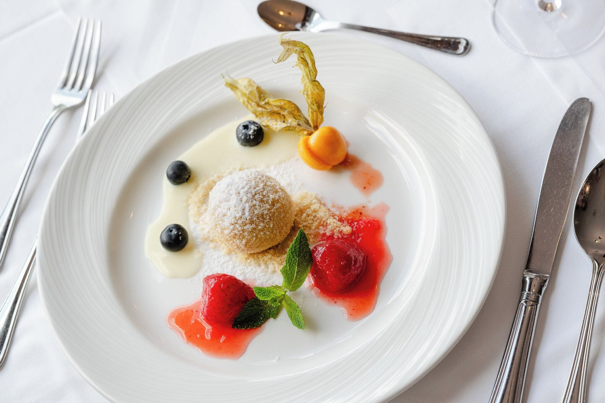 A white plate with a dessert consisting of a dusted pastry, blueberries, a cherry, and a mint leaf.