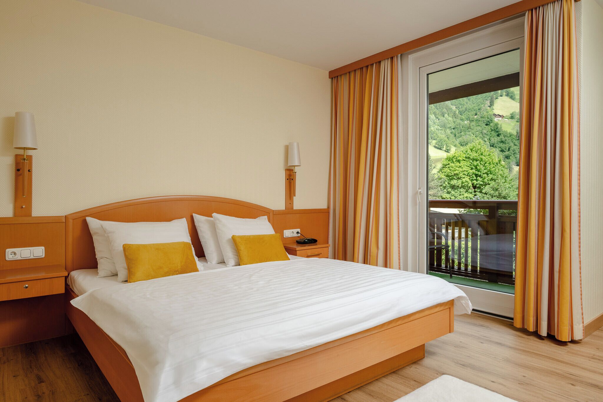 A hotel room with a large bed, a nightstand, a lamp, and a view of a balcony with curtains.
