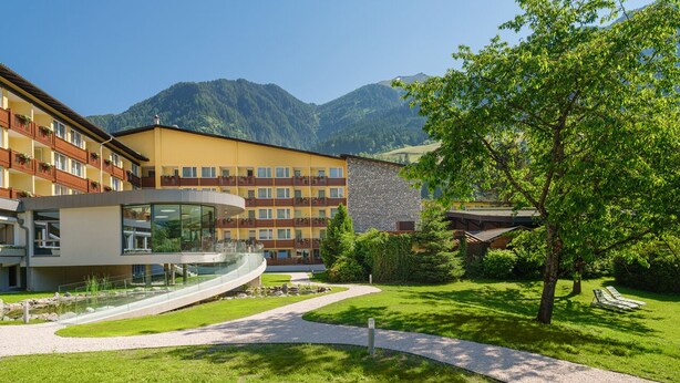 Un hotel con un edificio moderno, una pasarela de cristal curvada y un patio ajardinado con montañas al fondo.