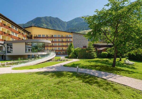 Un hotel con un edificio moderno, una pasarela de cristal curvada y un patio ajardinado con montañas al fondo.