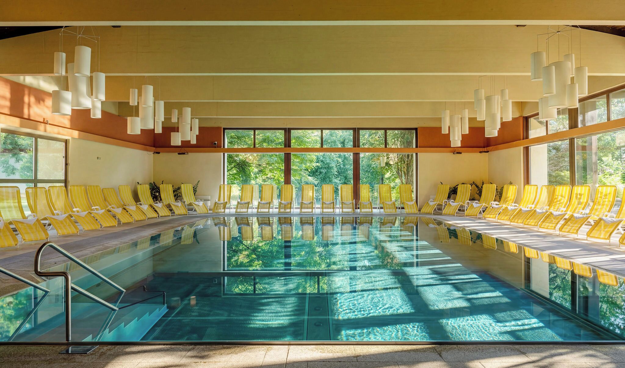 A large indoor swimming pool with multiple sun loungers arranged around it, large windows, and hanging pendant lights.