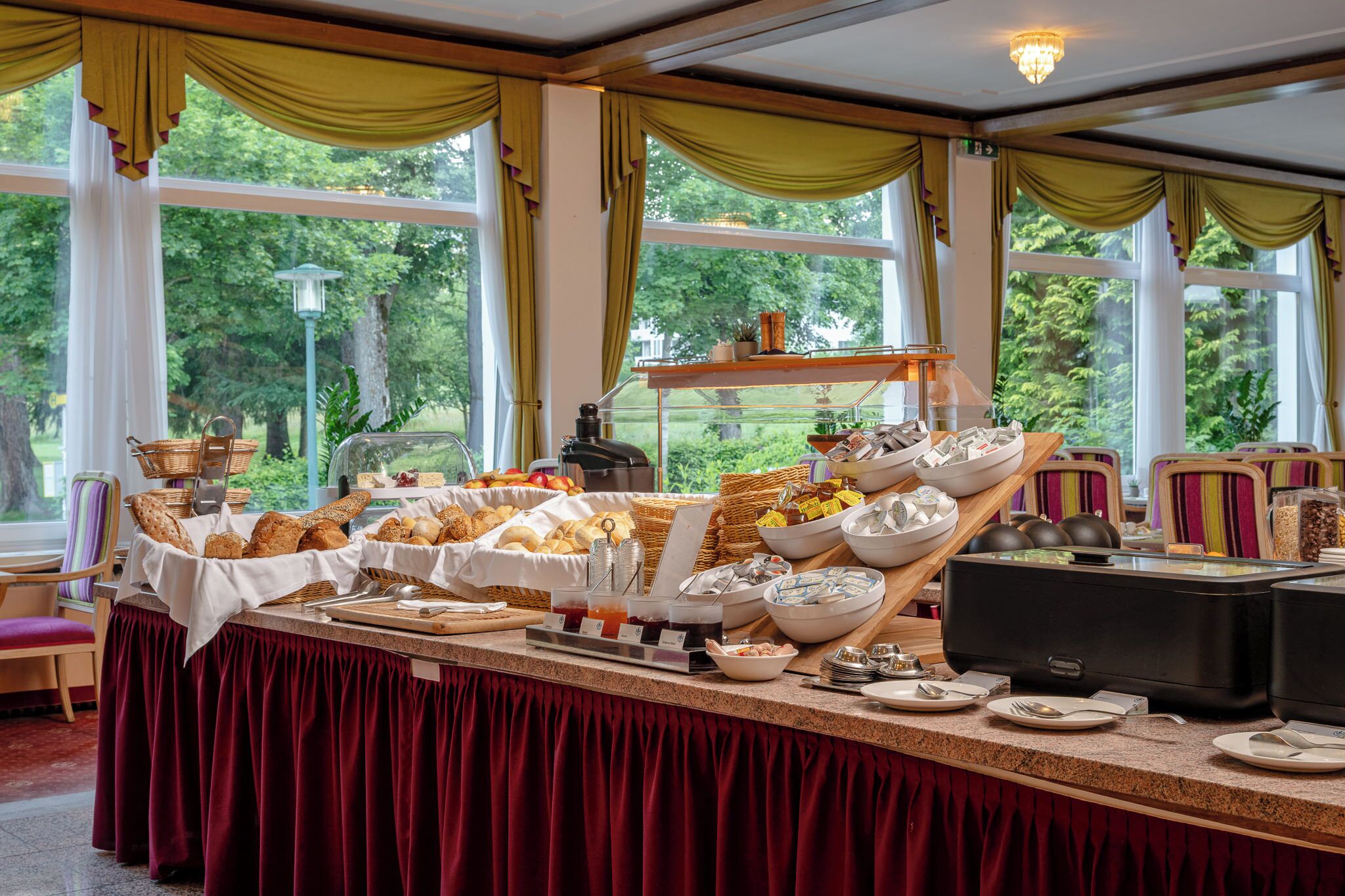 A buffet setup with various food items and a coffee station, set against a backdrop of large windows overlooking a green landscape.