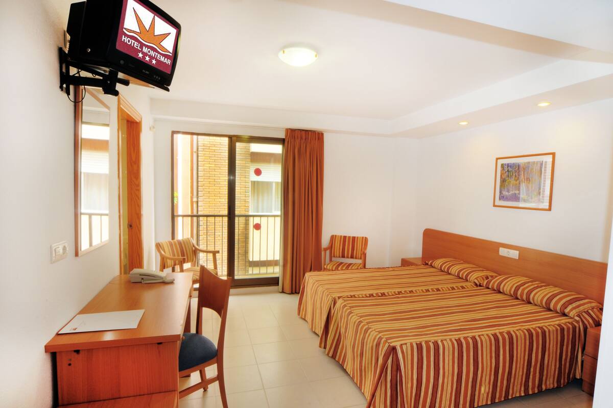 Habitación de hotel con escritorio, dos camas, televisor y balcón con vistas.