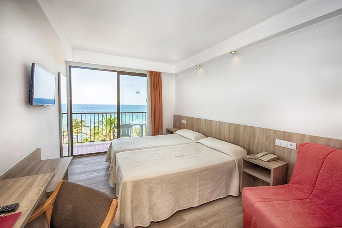 A hotel room with two beds, a desk, a chair, a TV, and a balcony with a view of the sea.
