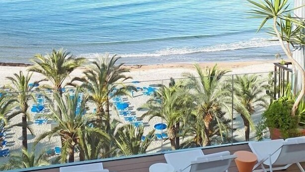 A balcony with white lounge chairs and a small table, overlooking a beach with palm trees and a pool. - Hotel Montemar Benidorm (Benidorm)