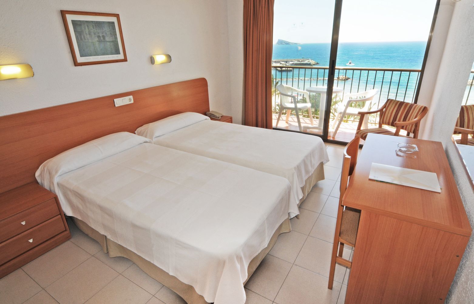 Habitación de hotel con dos camas, un escritorio y un balcón con vista al mar.