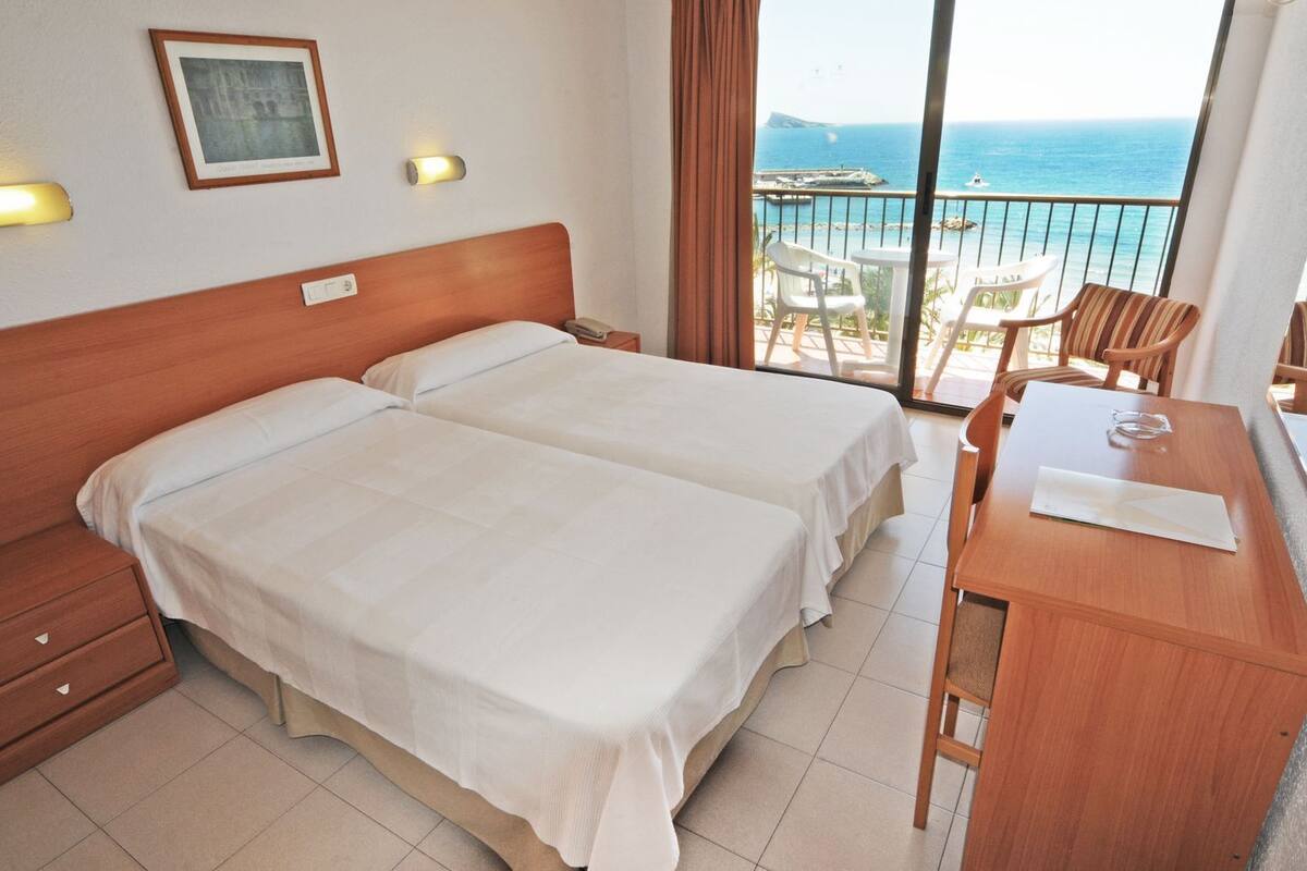 Habitación de hotel con dos camas, un escritorio y un balcón con vista al mar.