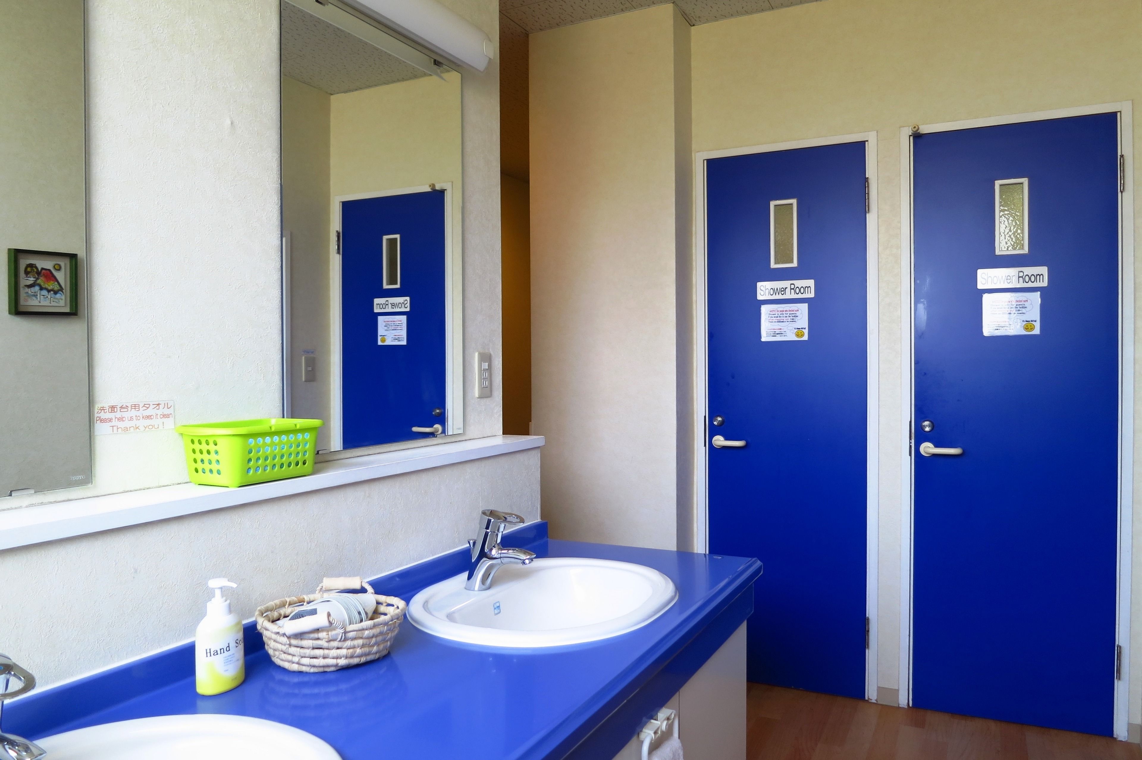 A bathroom with two blue doors labeled "Shower Room," a mirror, a sink with a soap dispenser, and a green basket on the counter.