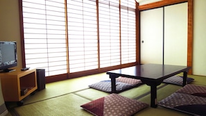 Um quarto japonês tradicional com tatames, uma mesa baixa, almofadas e uma televisão sobre um suporte de madeira.