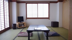 Une pièce avec un sol recouvert de tatamis, une petite table basse, des coussins, un téléviseur posé sur un meuble et une porte coulissante.