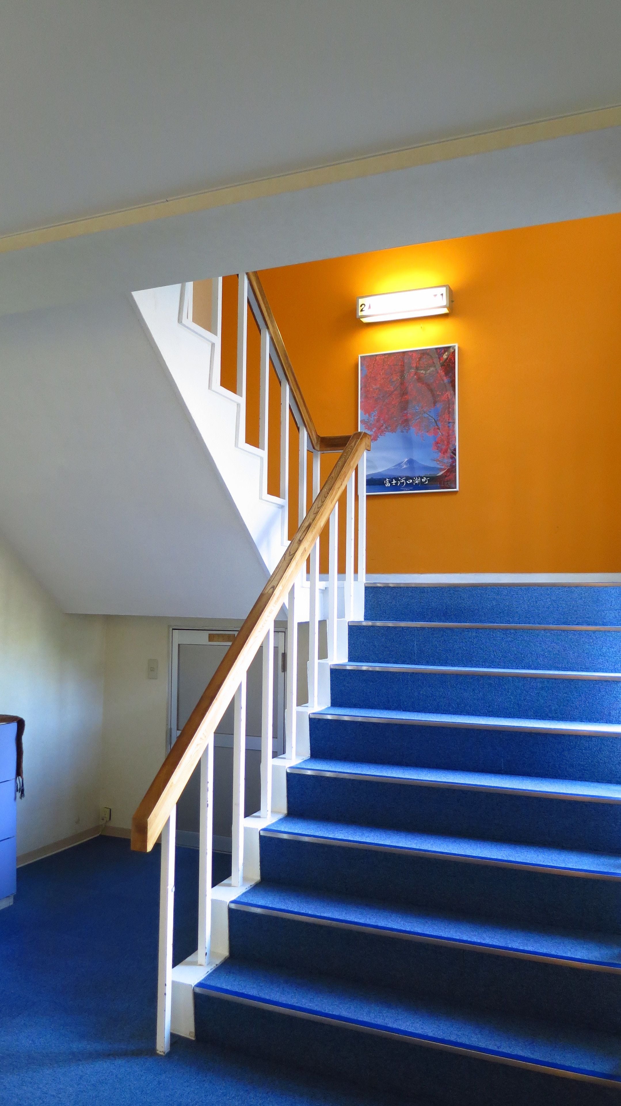 Eine Treppe mit blau beflochtenen Stufen und Holhandläufen, die zu einer Zwischenebene mit einer Leuchte und einem gerahmten Bild an der Wand führt.