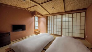 A traditional Japanese-style room with tatami flooring, two beds with white bedding, a sliding shoji screen, a television on the wall, and a small wooden stool.