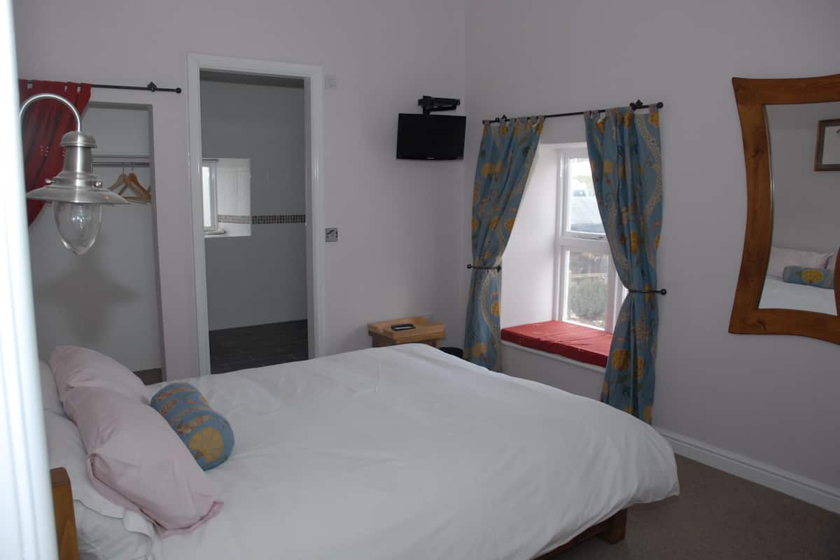 A bedroom with a bed, a window with curtains, a bedside table, a mirror, and a wall-mounted TV.