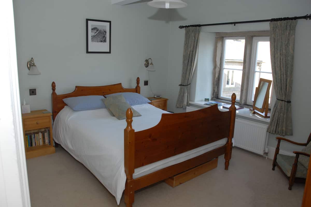 A bedroom with a wooden bed, bedside tables, a nightstand with books, a window with curtains, and a framed picture on the wall.