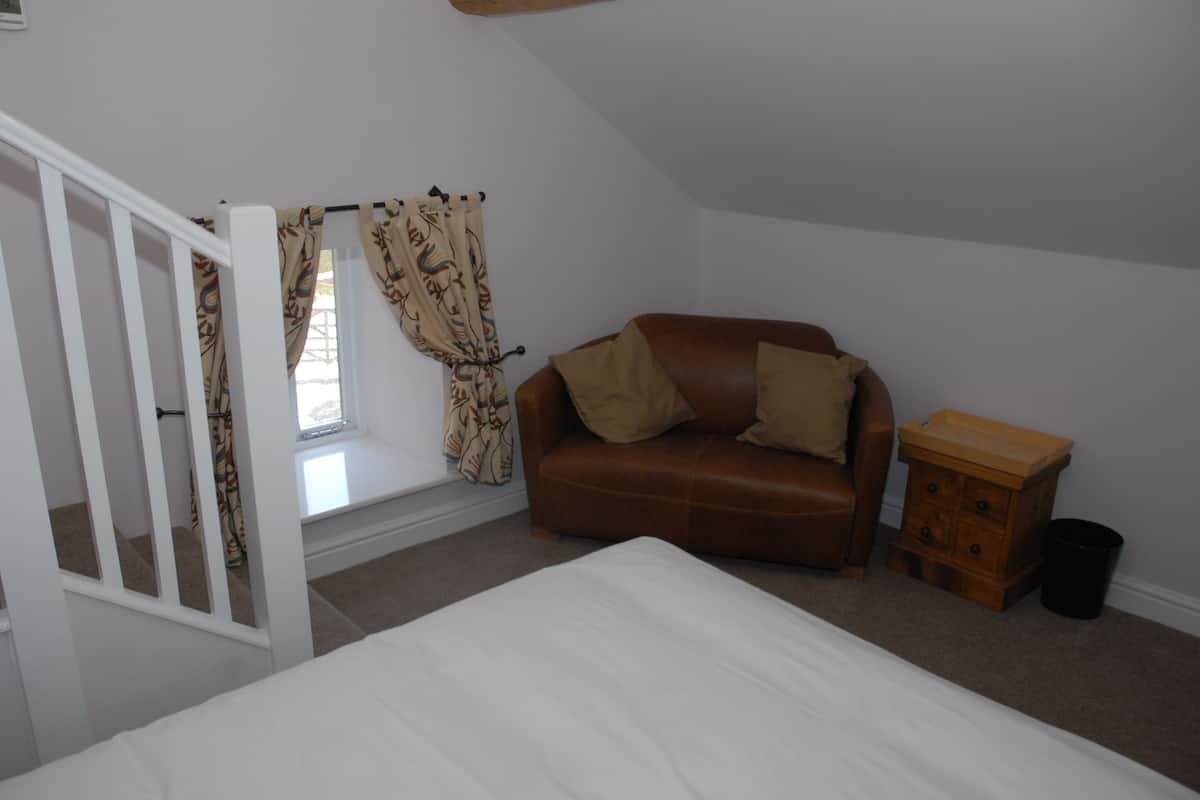 A bedroom with a bed, a brown leather sofa, a window with patterned curtains, a wooden nightstand, and a white staircase.