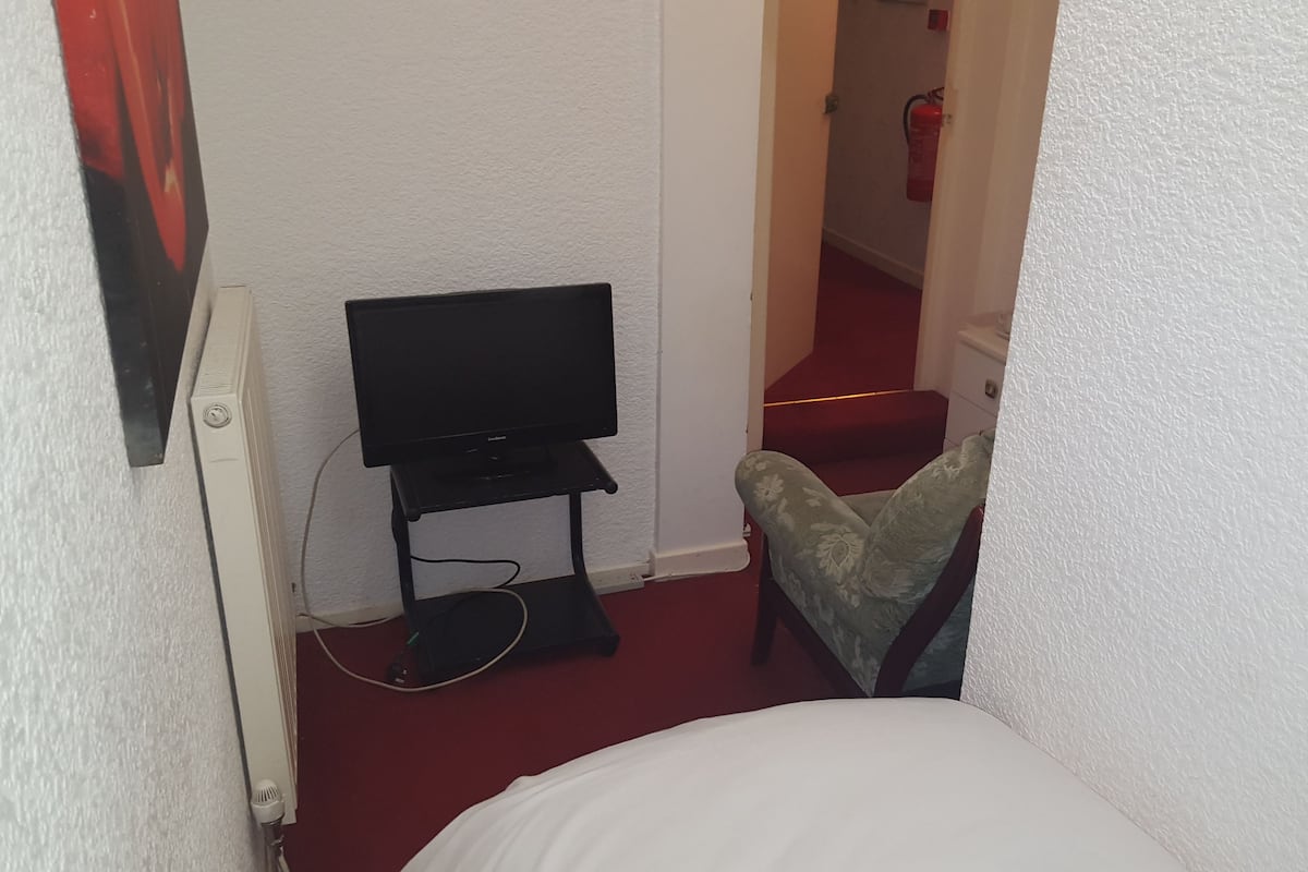 Ein kleines Zimmer mit einem weißen Bett, einem Fernseher auf einem Schrank und einem Sessel.
