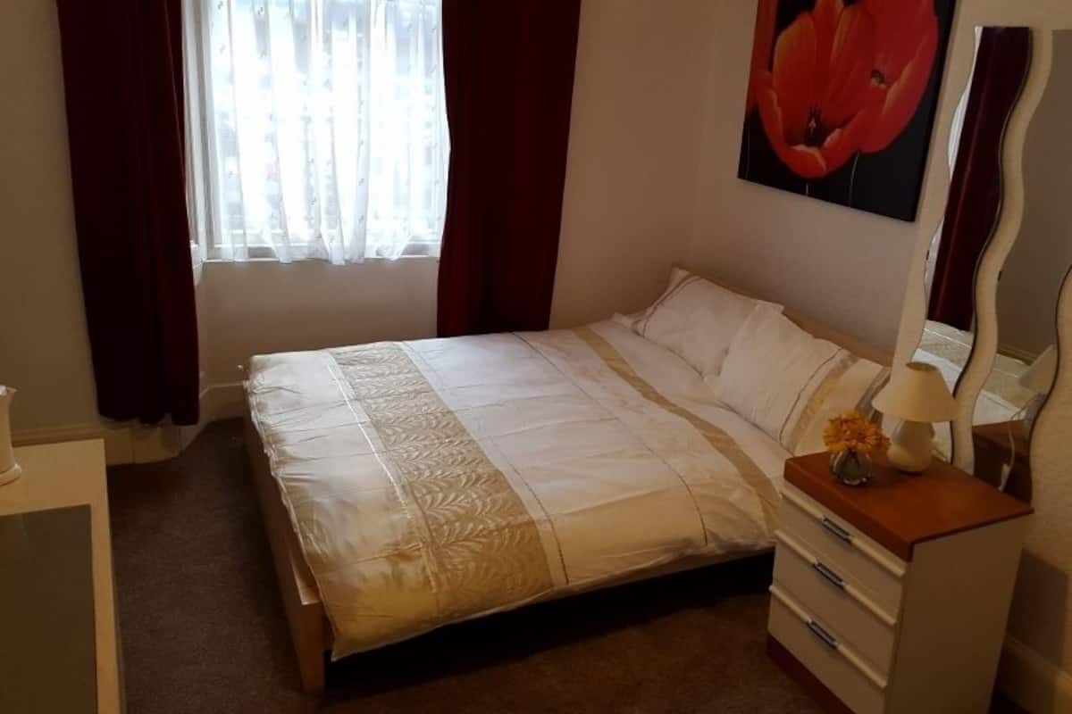 Ein ordentlich bezogenes Bett mit weißer und beiger Bettdecke, ein Nachttisch aus Holz mit Lampe und Vase sowie ein großer Spiegel an der Wand.