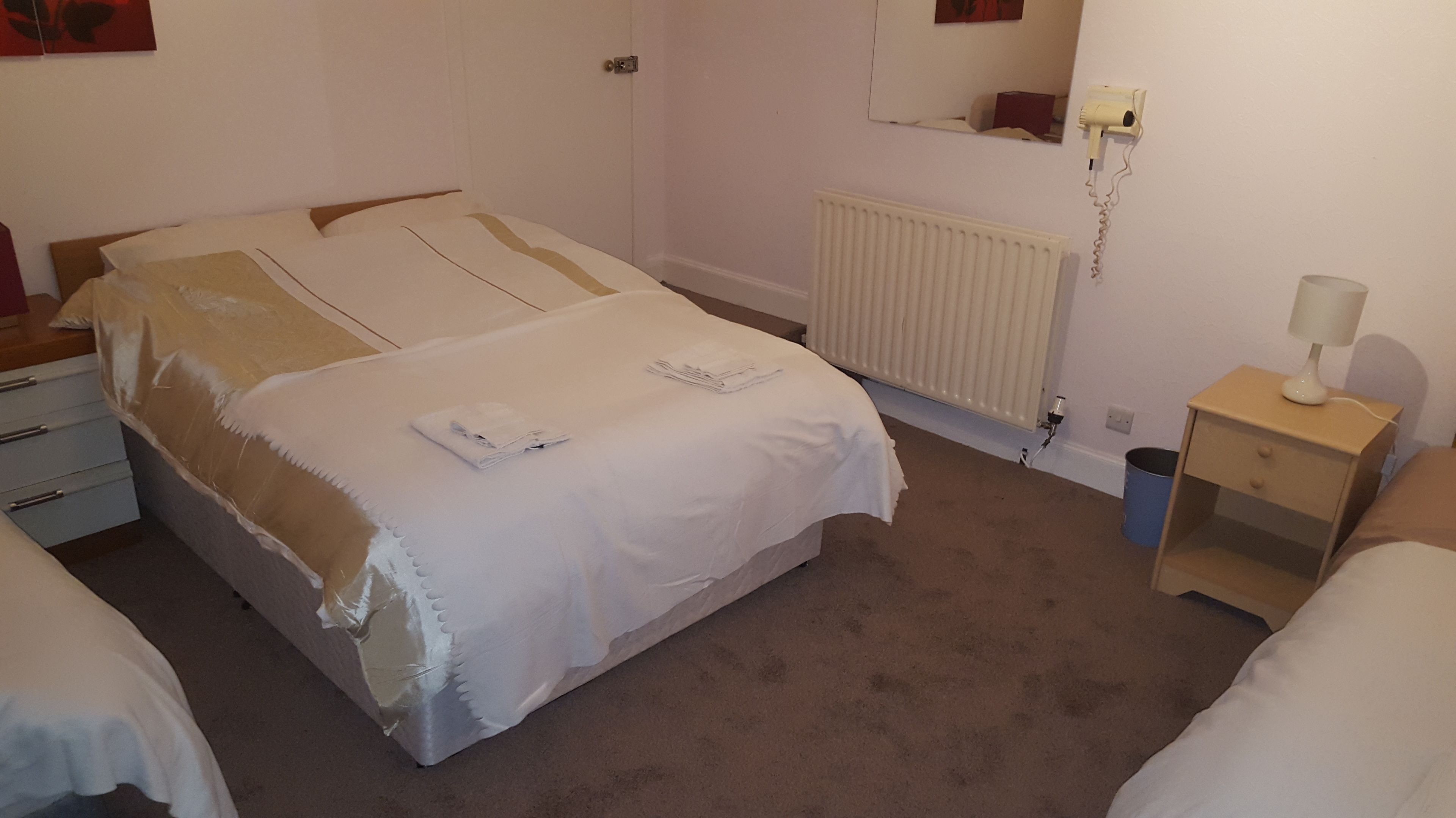 Un dormitorio con cama, mesita de noche, radiador y secador de pelo fijado a la pared.