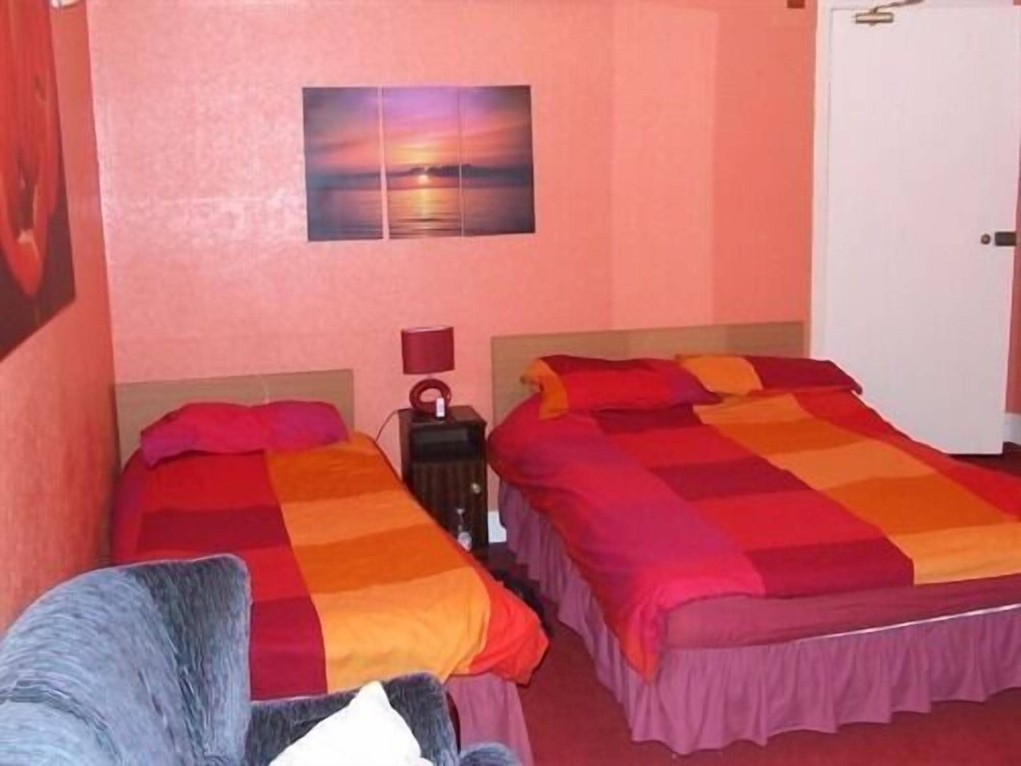 Una habitación con dos camas, cada una con una colcha rojo anaranjada, una mesita de noche con lámpara y un cuadro colgado en la pared que muestra un atardecer.