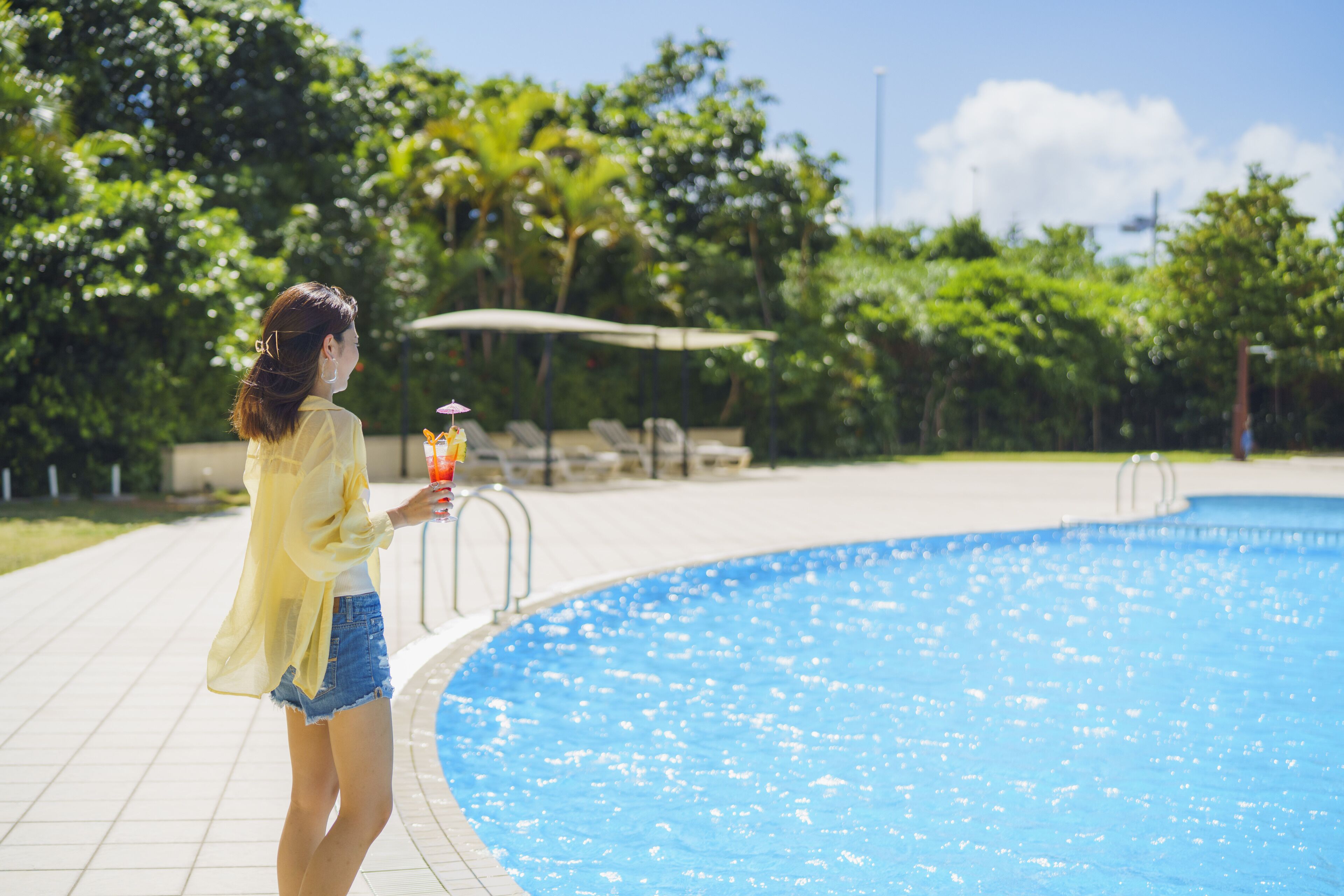 A person standing by a swimming pool with a clear sky and greenery in the background.