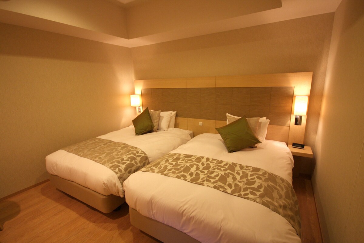Habitación de hotel con dos camas, cada una con cabecera, lámparas de noche y colcha estampada.
