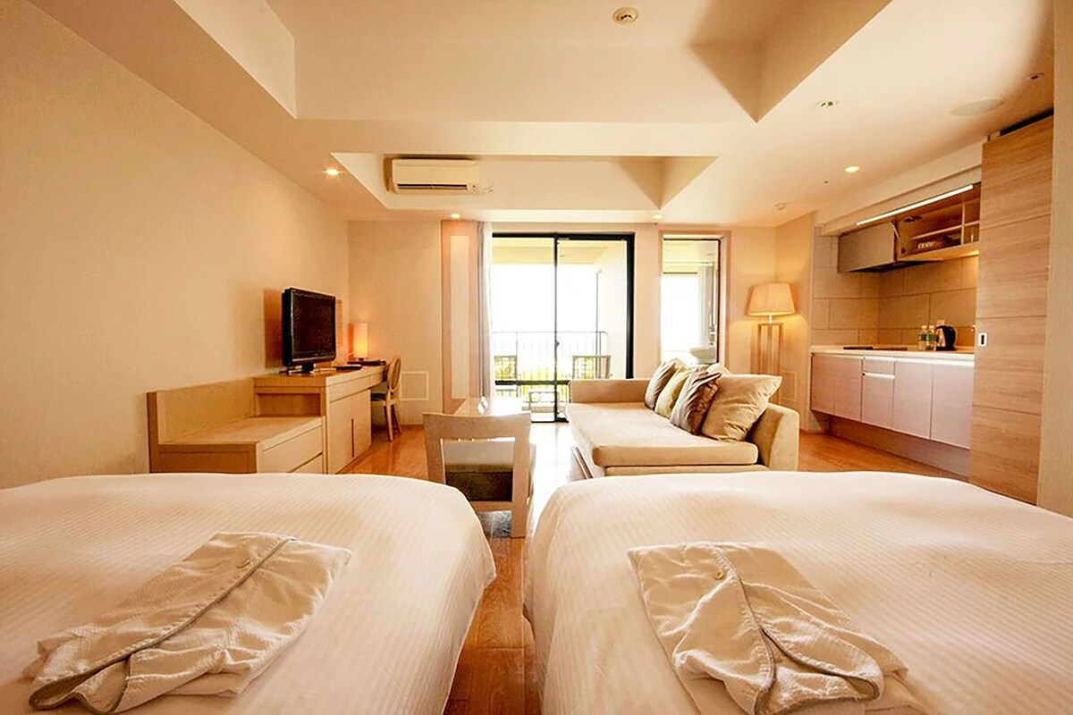 Habitación de hotel con dos camas, televisión, escritorio, silla, sofá, balcón y kitchenette.