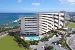 A multi-story hotel with a swimming pool, parking area, and a view of the ocean.