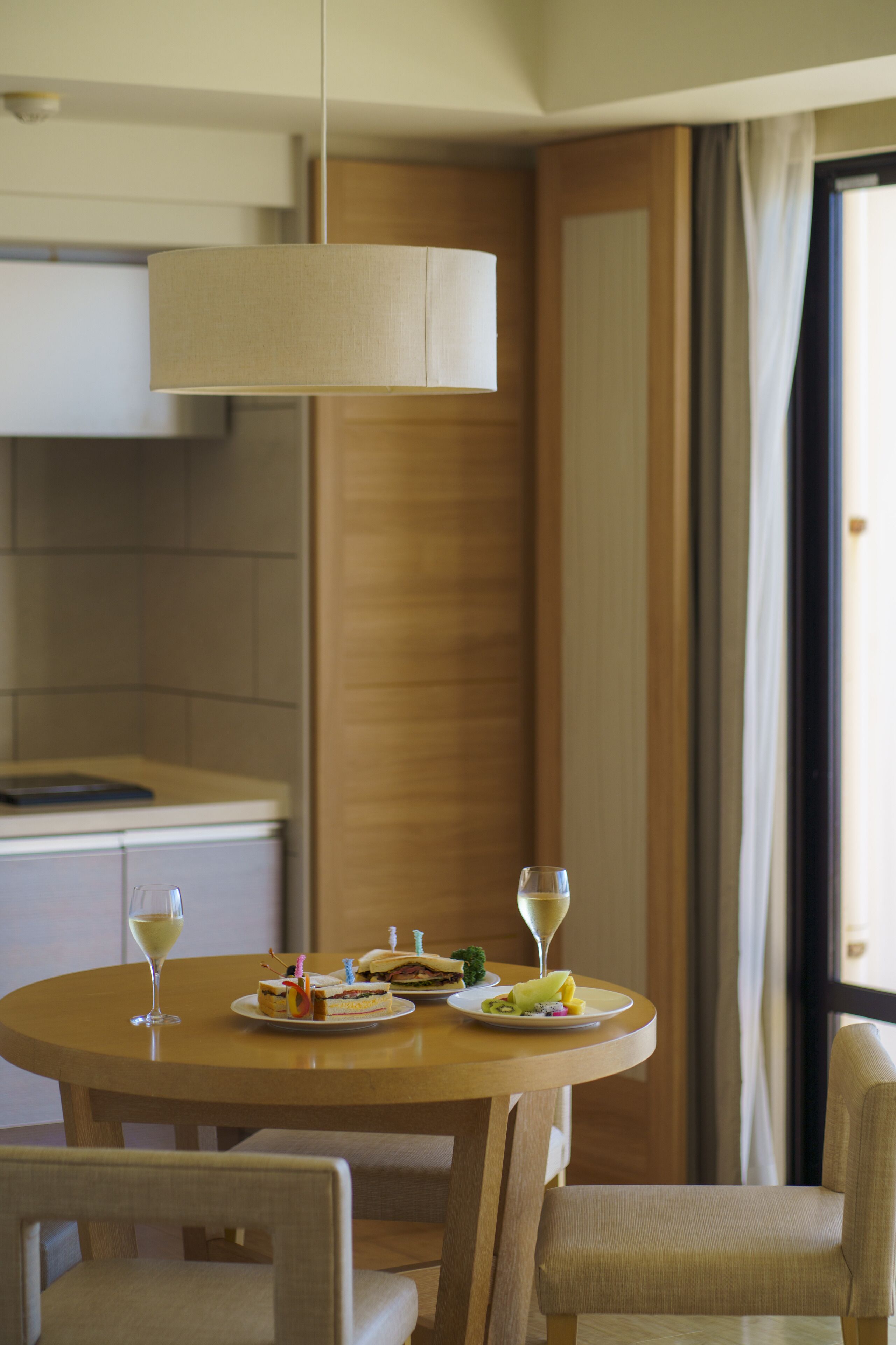 A dining area with a round table, two chairs, a hanging light fixture, and a view of the outdoors through a window.