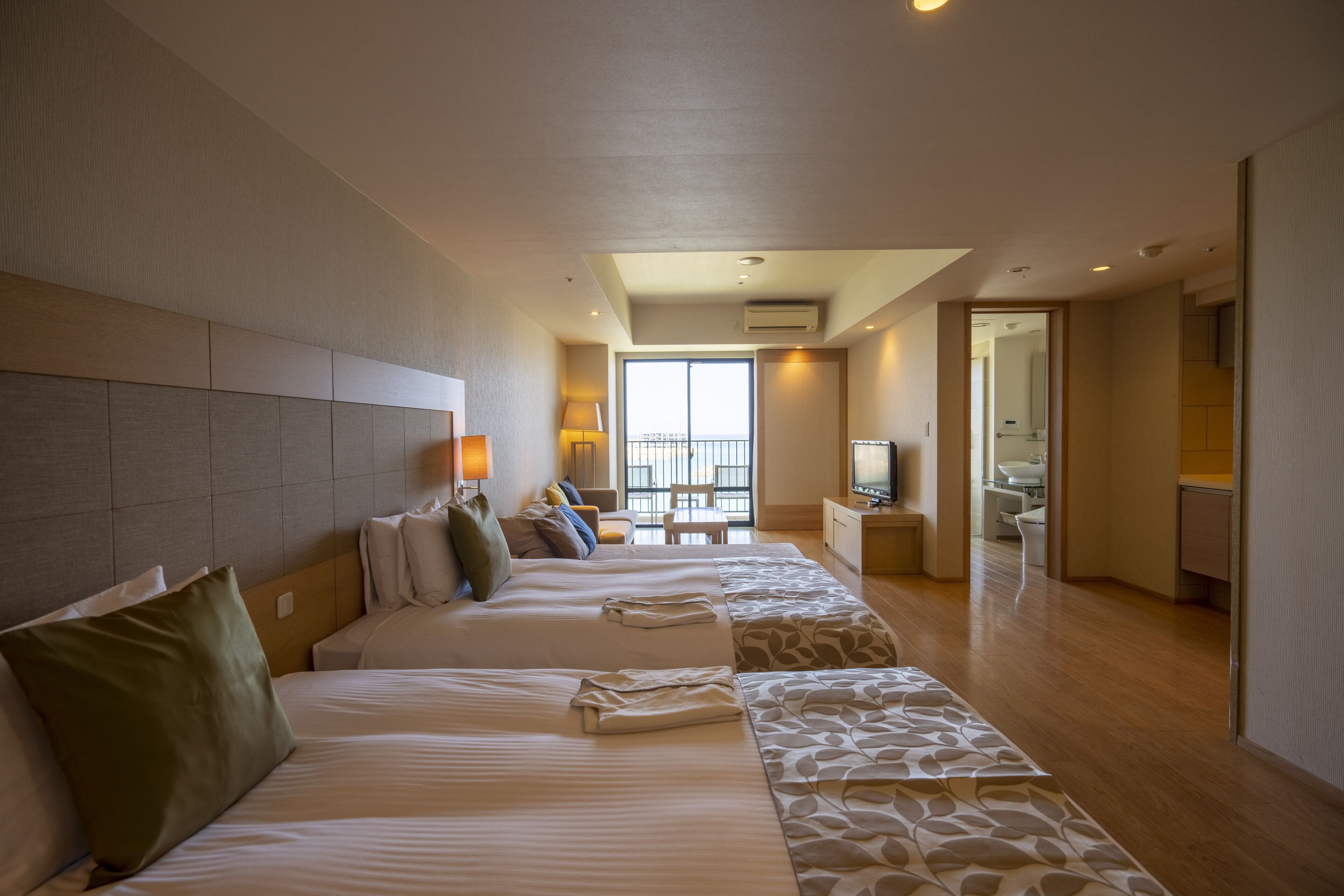 A hotel room with a large bed, a smaller bed, a sofa, a desk, and a bathroom visible through an open door.
