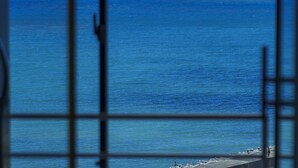 Vista a través de una ventana con marco metálico, que da a una playa con agua tranquila y cielo despejado.