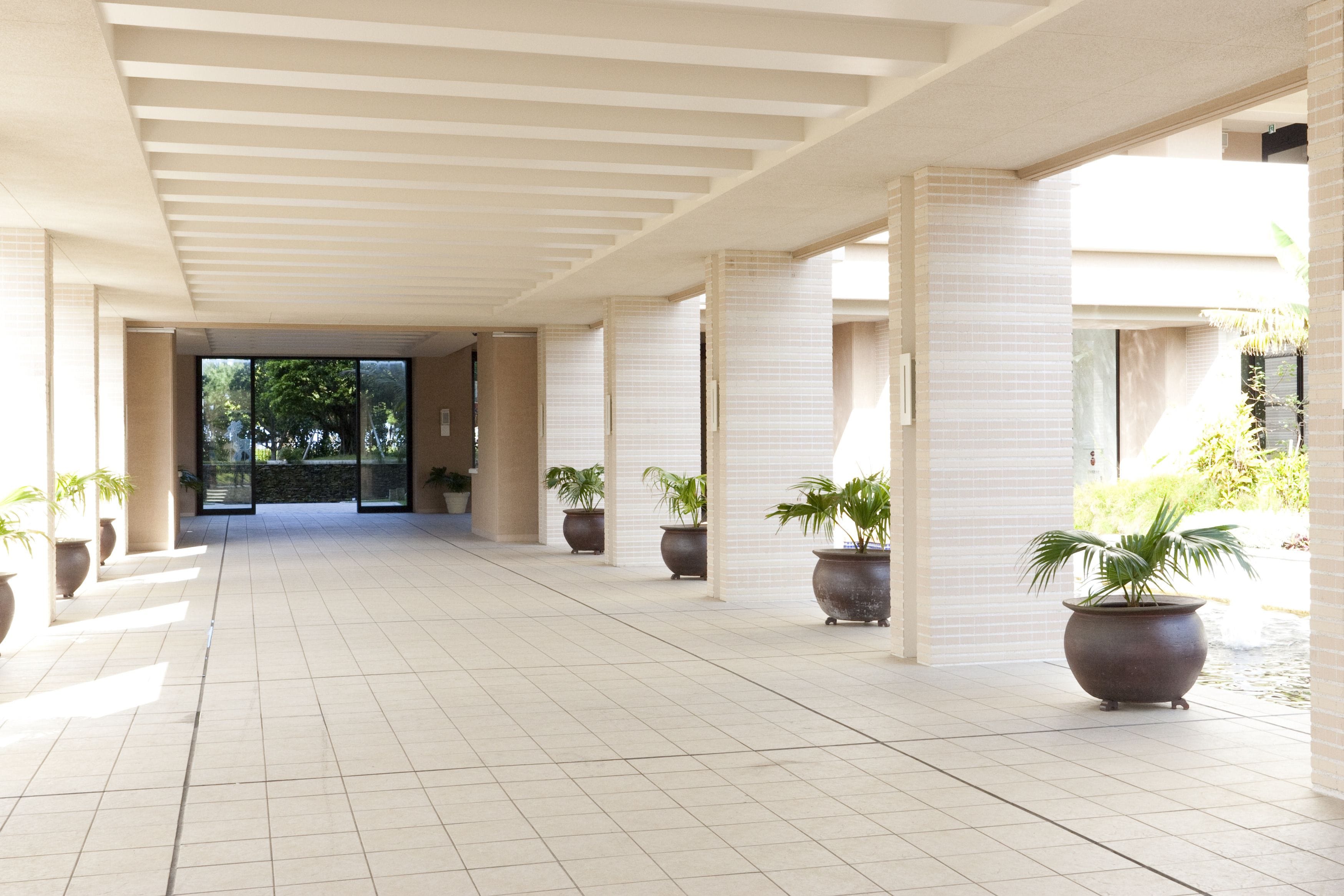 A spacious corridor with tiled flooring, large potted plants, and a modern architectural design.