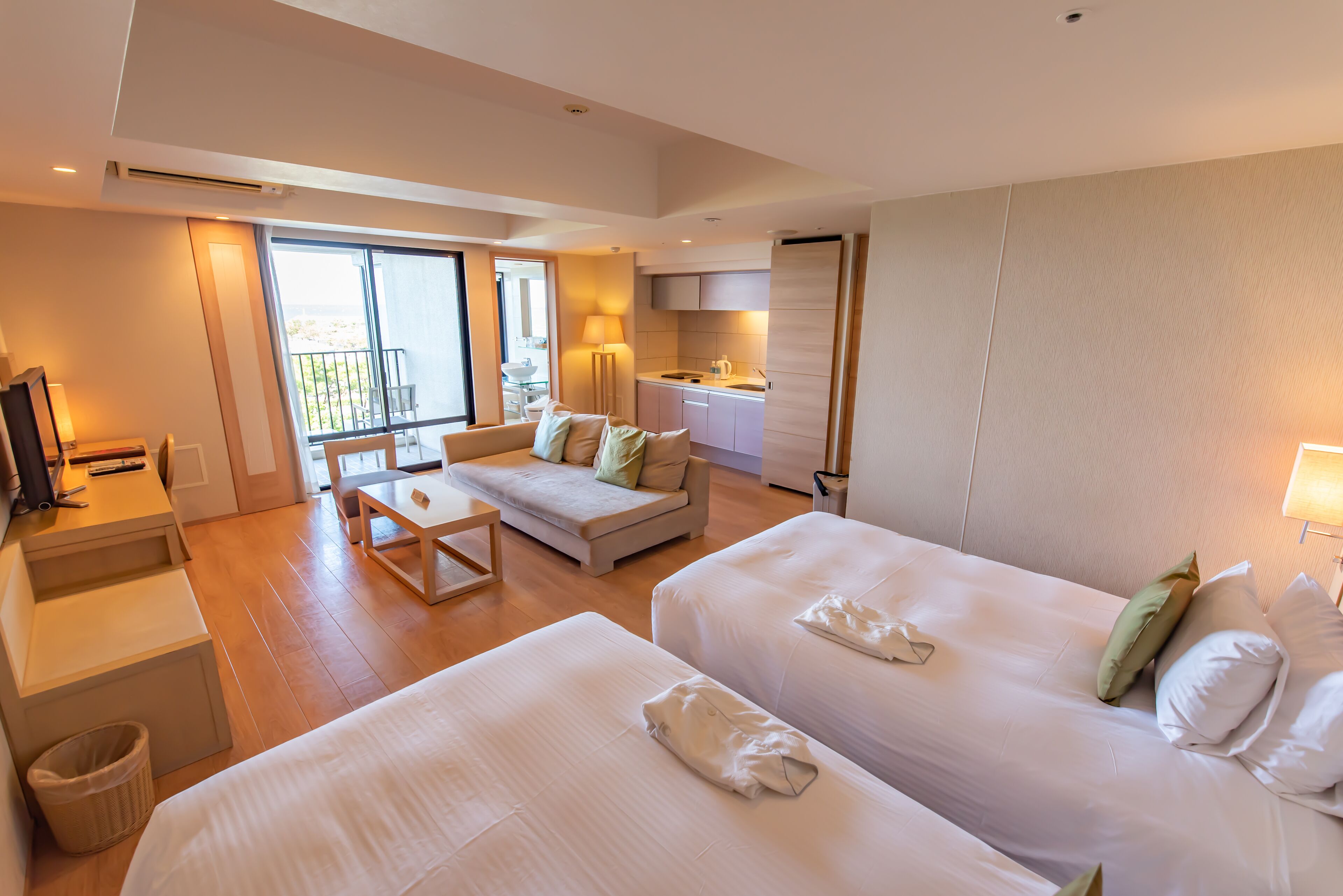 A hotel room with two beds, a sofa, a small table, a desk, a kitchen area, and a balcony with a view.