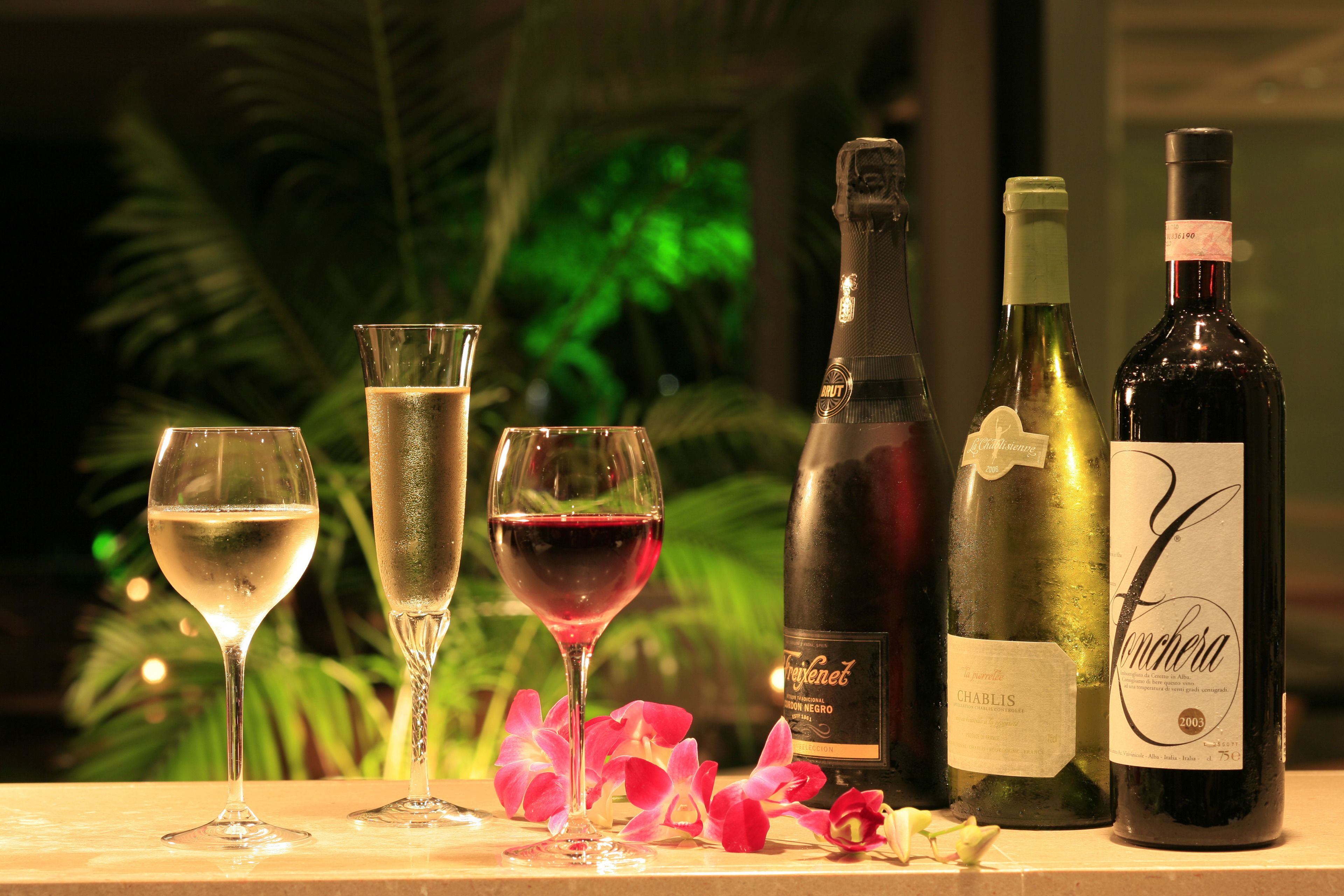 A table with four glasses of wine and three bottles of wine, surrounded by pink flowers.