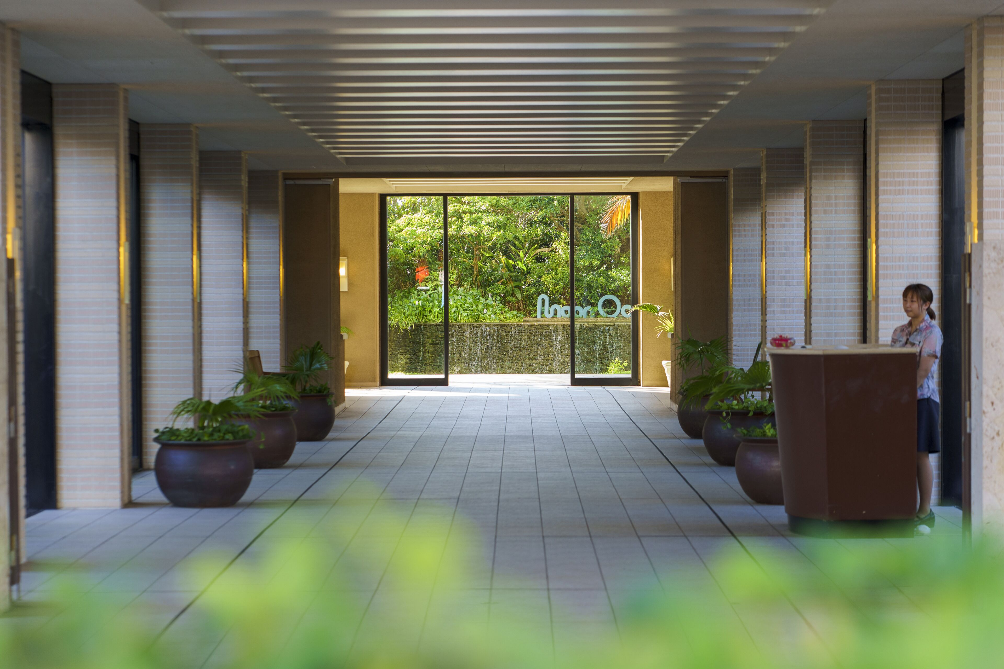 A hotel lobby with a reception desk, potted plants, and a large glass door leading to a garden area.