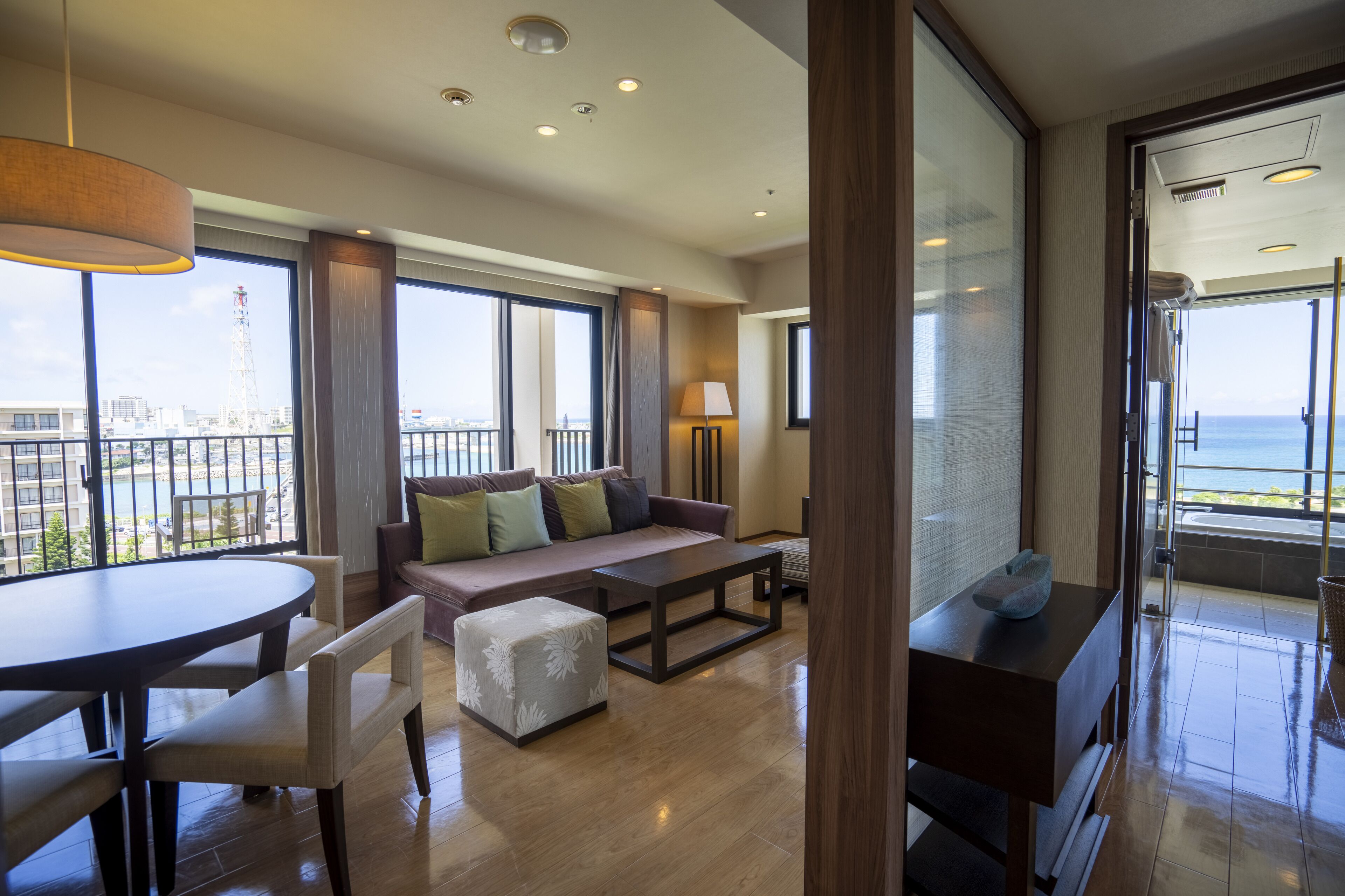 Una sala de estar moderna con un sofá, reposapiés y una mesa de centro, ventanas amplias con vista al mar, y un balcón con barandilla.