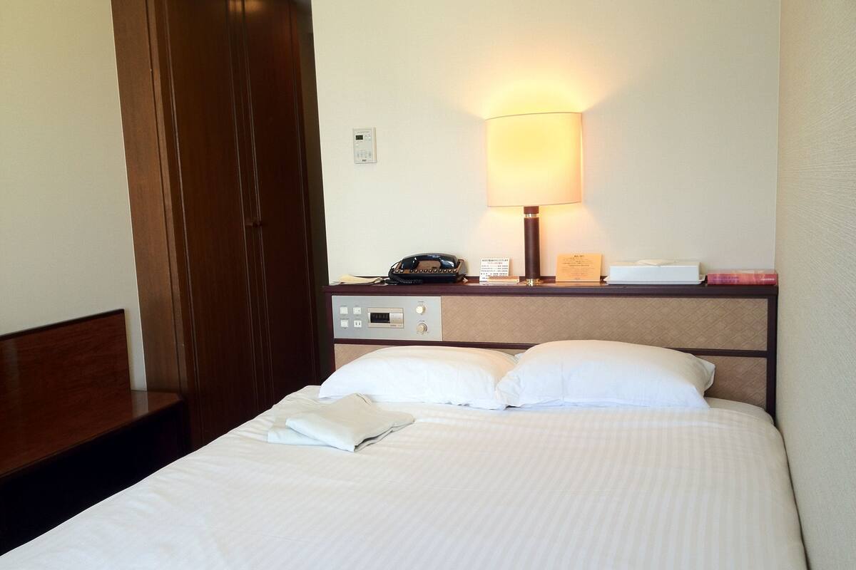 A hotel room with a bed, a nightstand, a lamp, a phone, and a control panel.