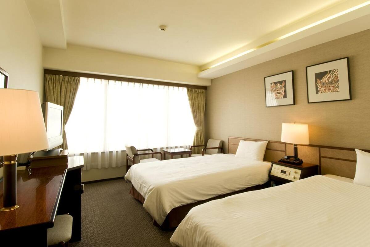 A hotel room with two beds, a desk, a chair, a TV, and a window with curtains.