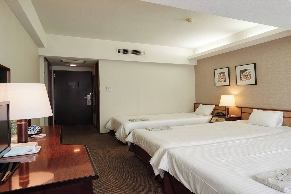 A hotel room with two beds, a desk with a lamp, and a door leading to another room.