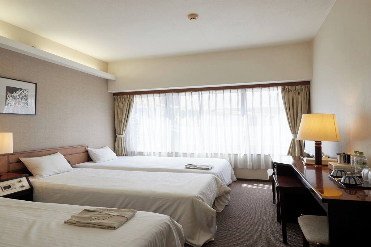 A hotel room with two beds, a desk, a chair, a lamp, a mirror, and a window with curtains.