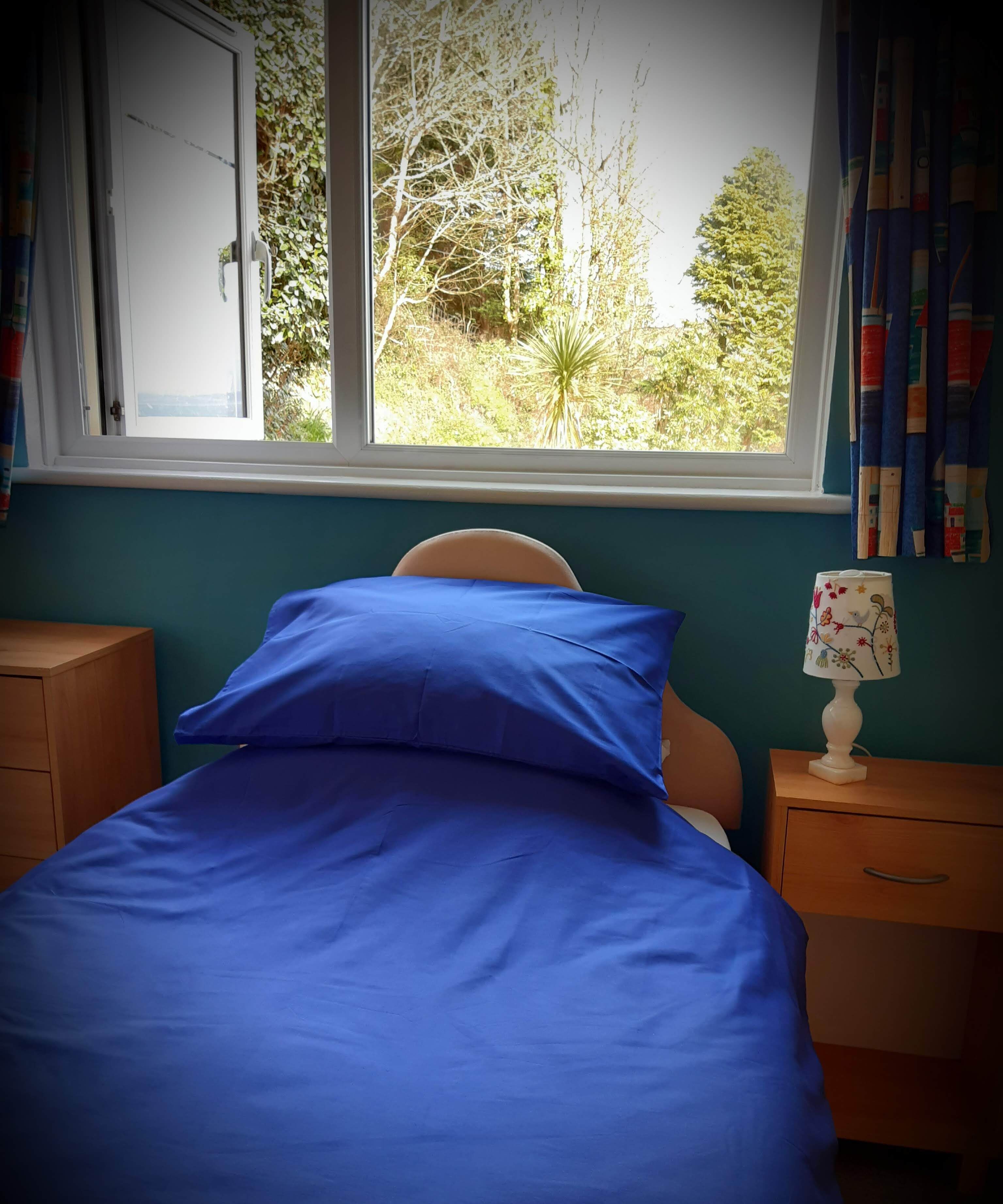 Quarto com cama, mesa de cabeceira, luminária e janela com vista para árvores e vegetação.