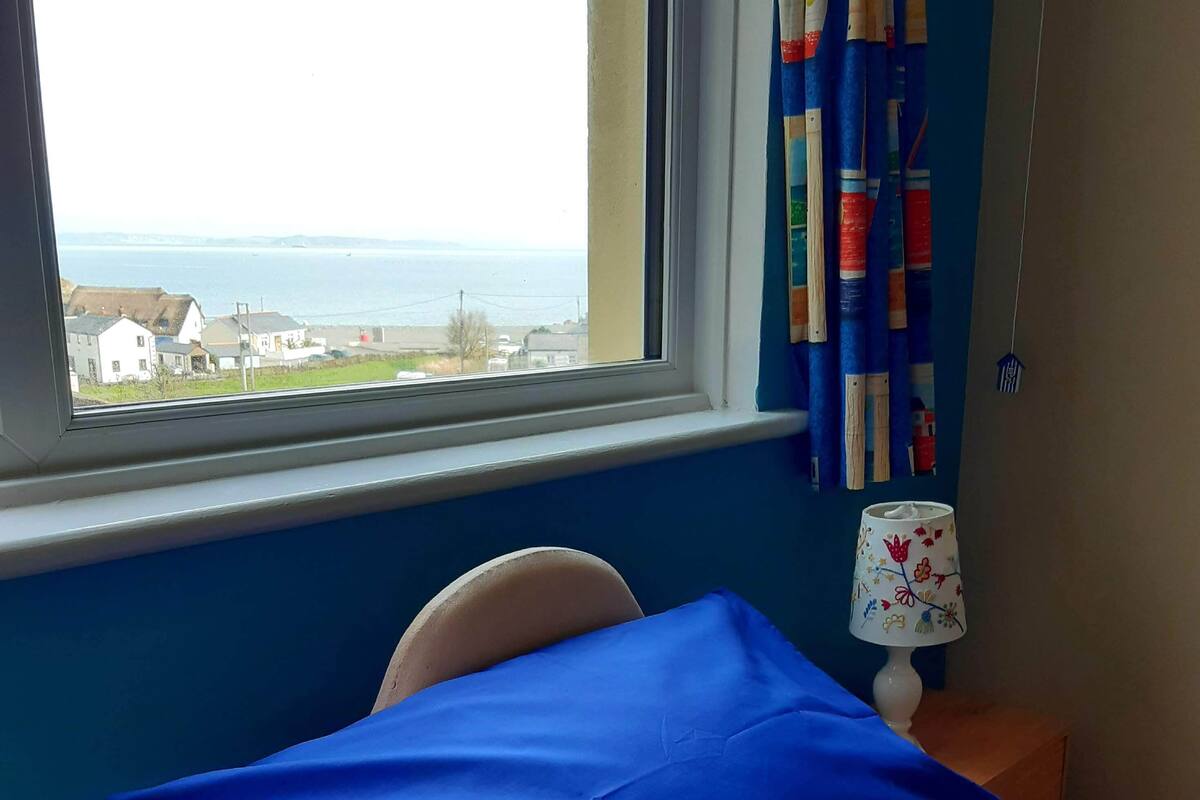 A bedroom with a bed, a window with a view of houses and the sea, a lamp, and a curtain.