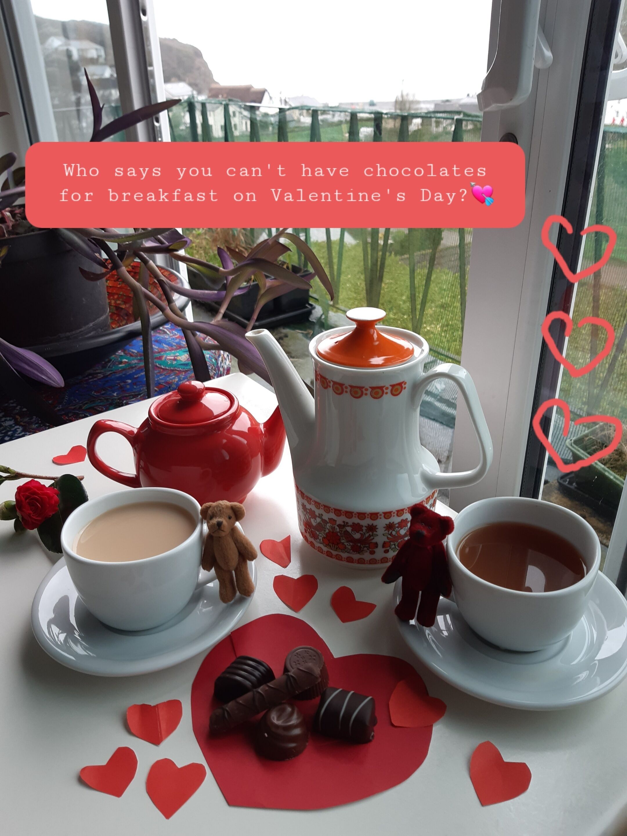 A breakfast setup with a teapot, cups, and chocolates on a table by a window.