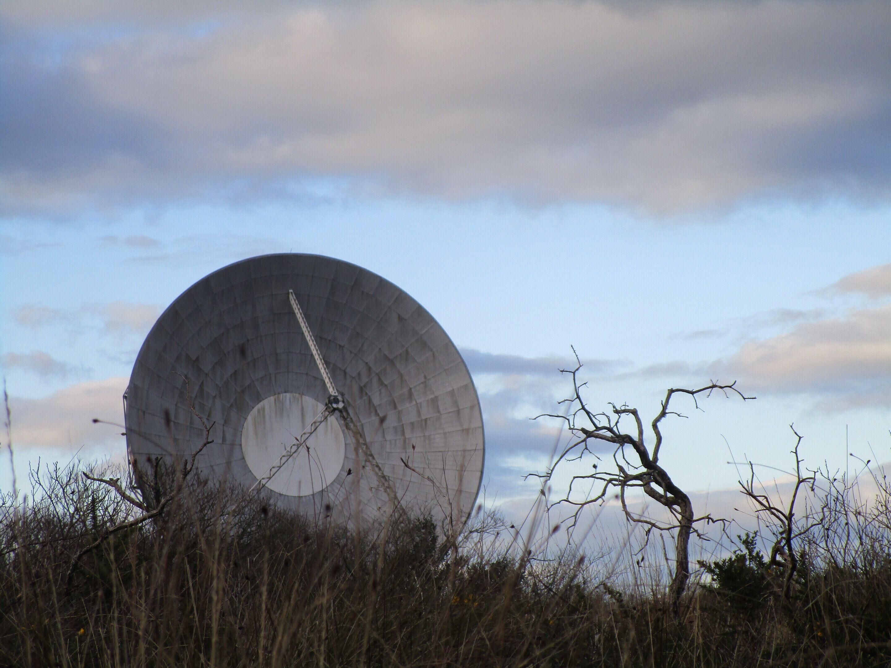 A large, circular structure with a central opening, surrounded by dry vegetation and a cloudy sky.