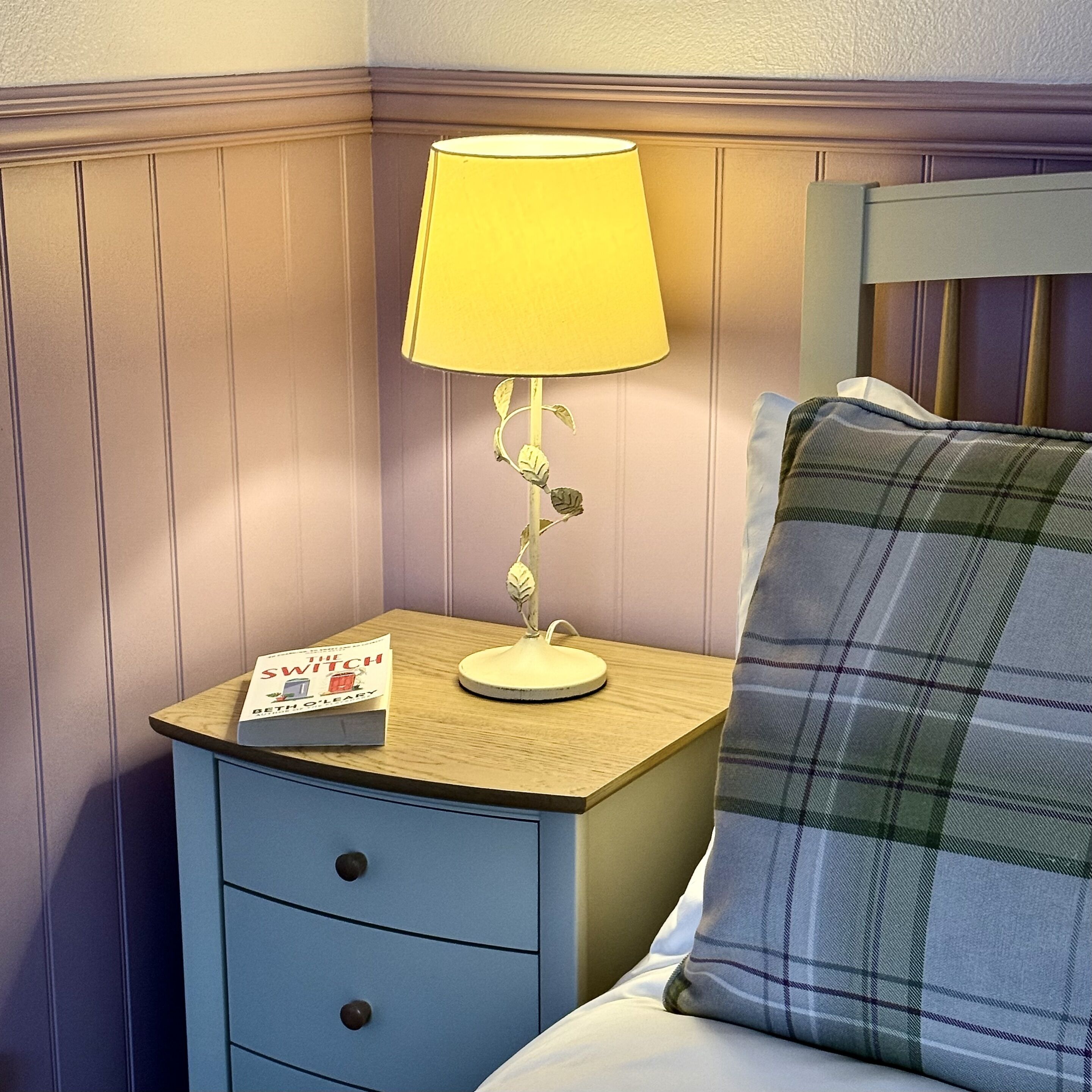 A neatly made bed with a plaid blanket, a bedside lamp, and a book titled "The Switch" on a wooden nightstand.